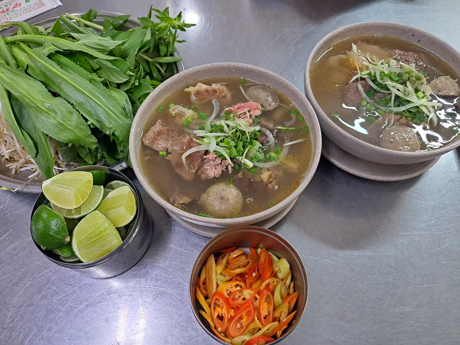 Fat med thaibasilika, ngo gai och böngroddar bredvid skålar med pho, lime och chili i Ho Chi Minh-staden