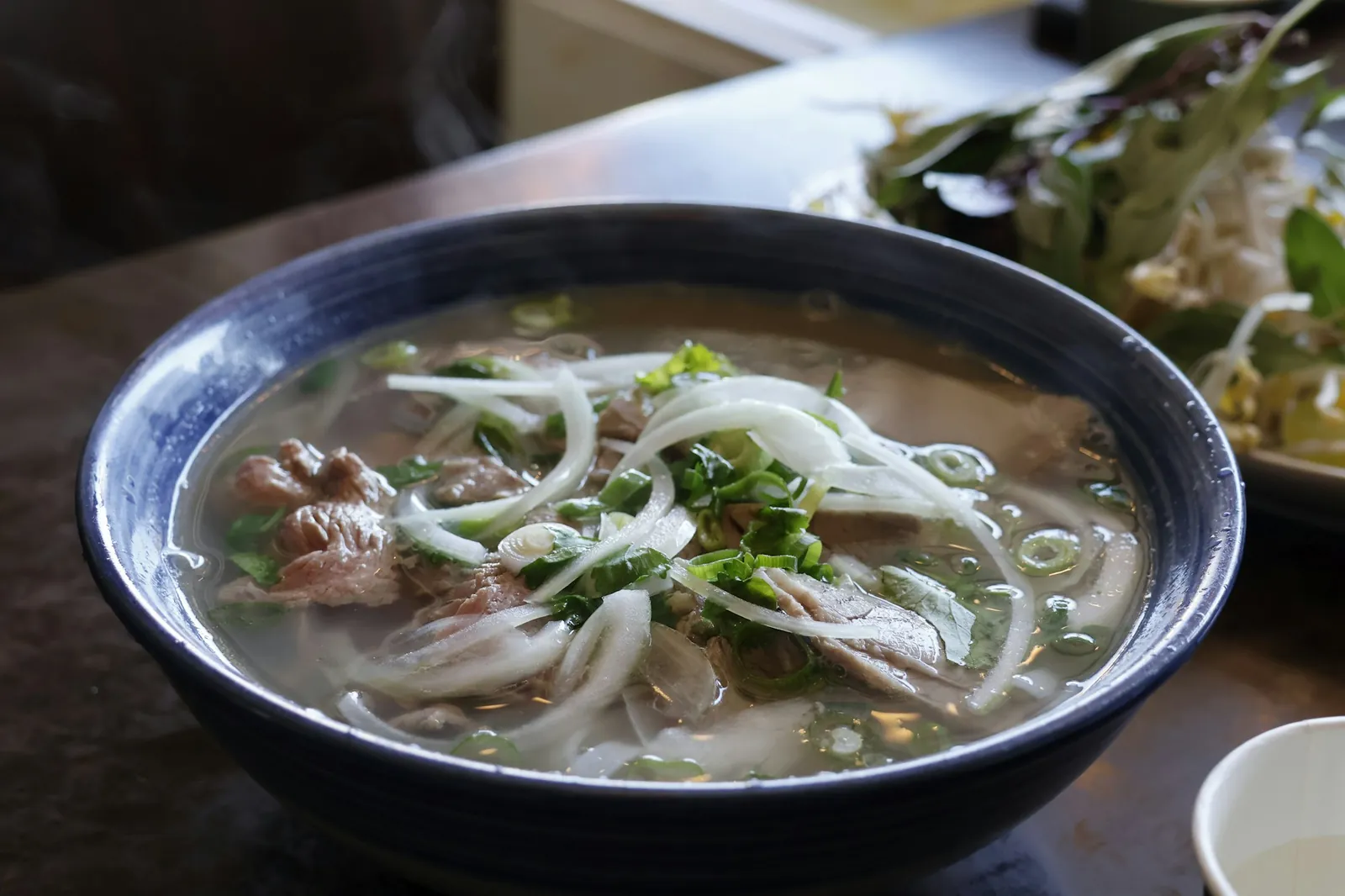 Skål med pho med tunna skivor nötkött, nudlar, lök och vårlök i klar buljong
