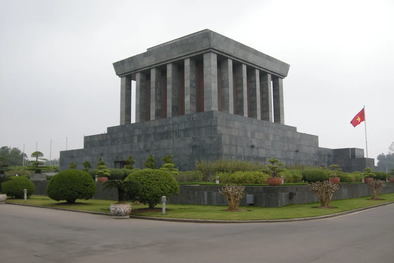 Ho Chi Minh-mausoleet i grå granit med prydnadsbuskar och Vietnams flagga i Ba Dinh, Hanoi