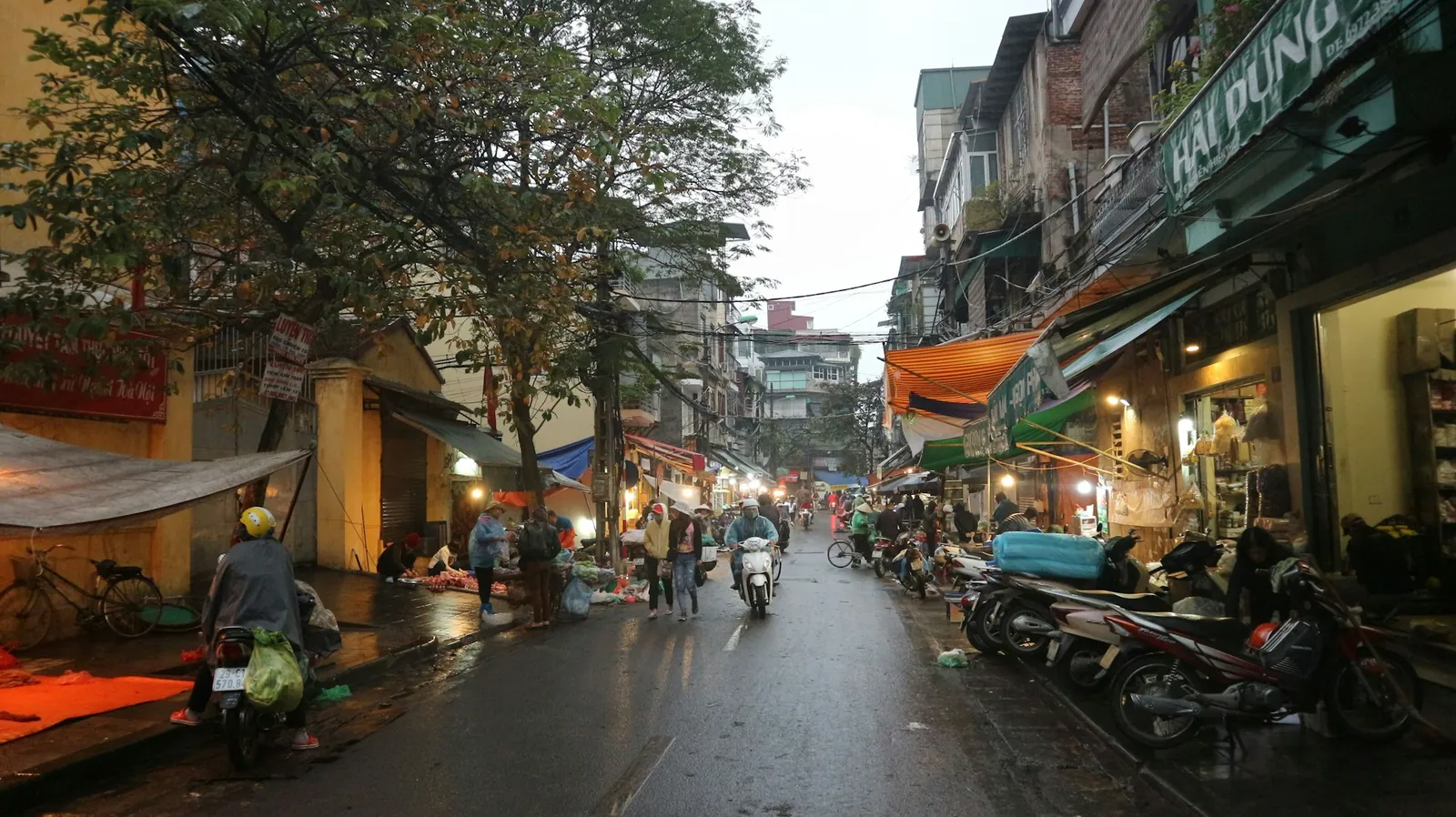 Smal gata med butiker, gatuförsäljare och mopeder i centrala Hanoi en regnig kväll