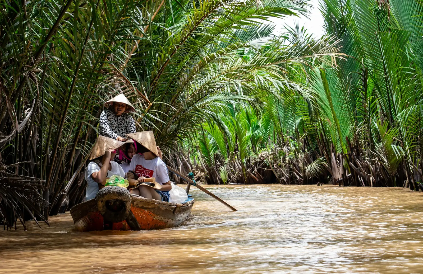 Kvinna i konisk hatt ror en liten båt genom en kanal kantad av vattenpalmer i Mekongdeltat