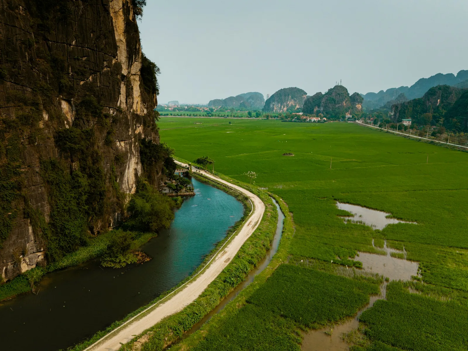 Gröna risfält, en kanal och en grusväg vid foten av kalkstensklippor i Ninh Binh