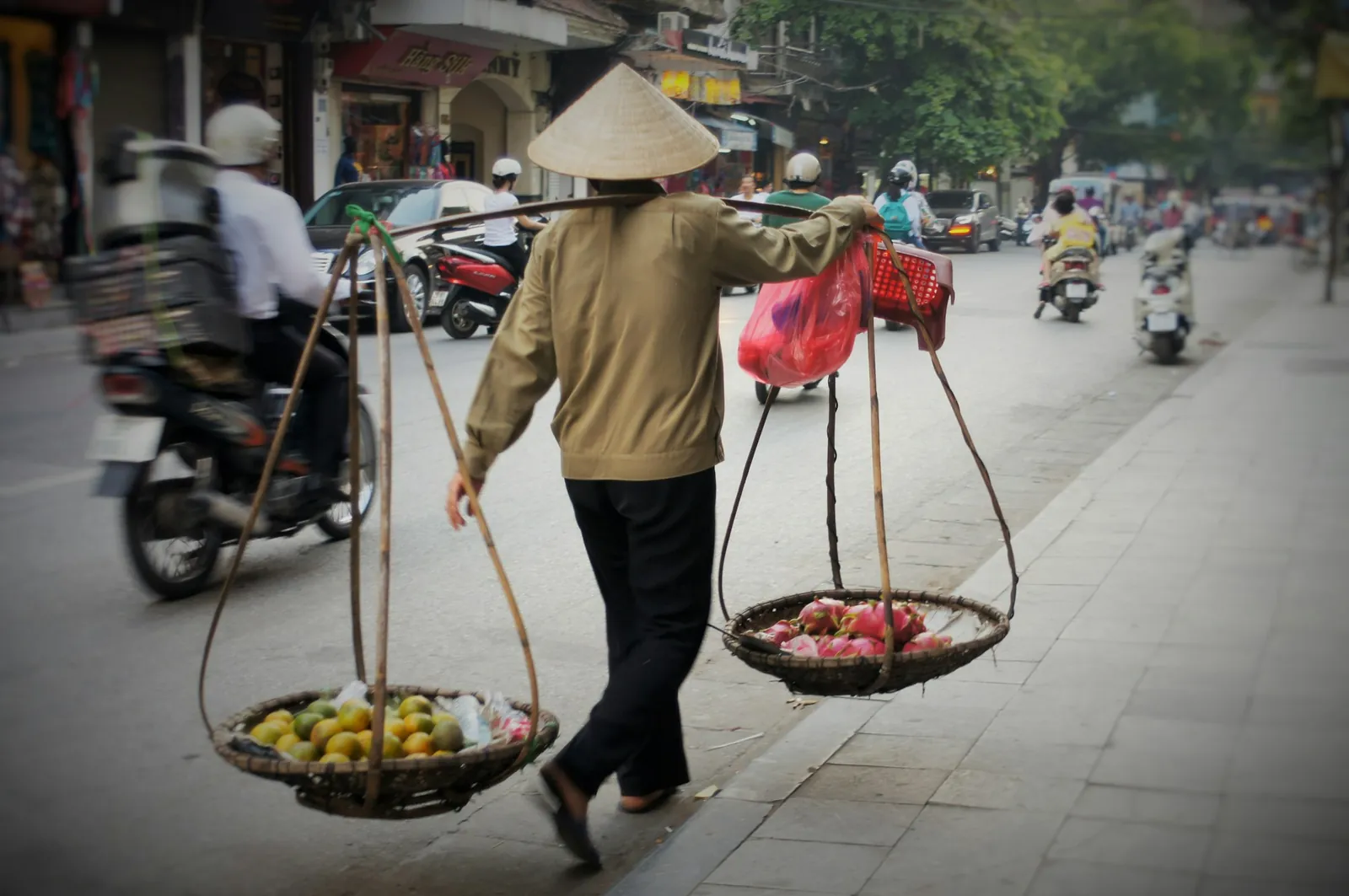 Gatuförsäljare i bladhatt med bärok och korgar med frukt på en stadsgata i Vietnam