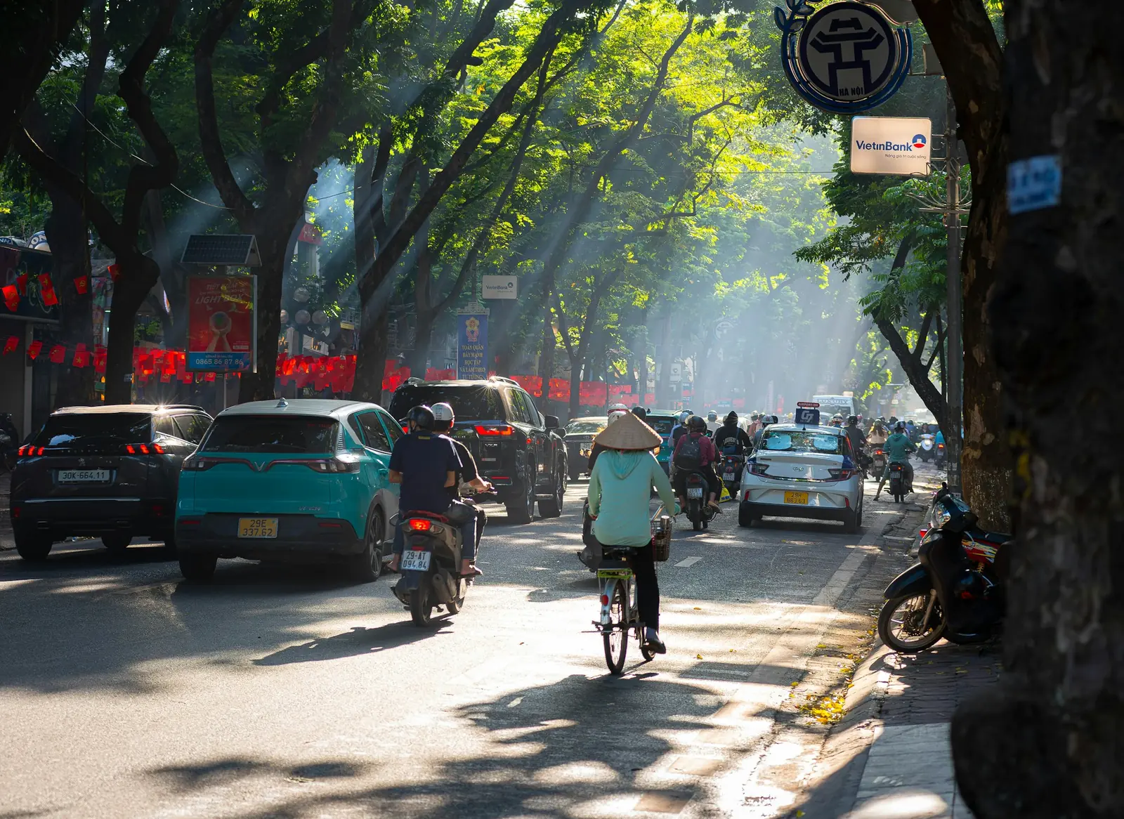 Starkt solljus silar genom träden över en trafikerad gata i Hanoi, en cyklist i bambuhatt