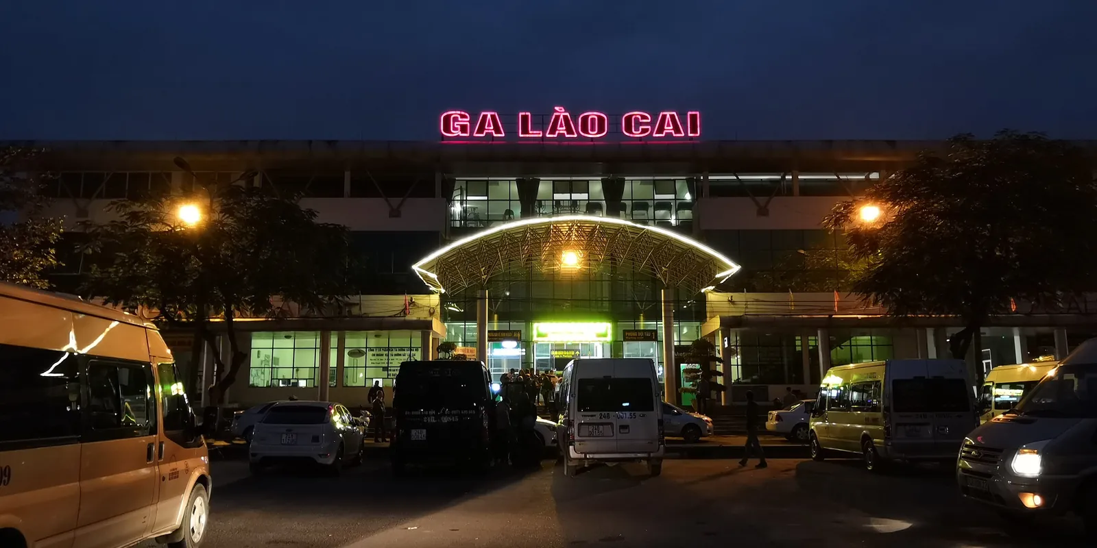 Lao Cais järnvägsstation i skymningen med upplyst skylt och minibussar framför entrén